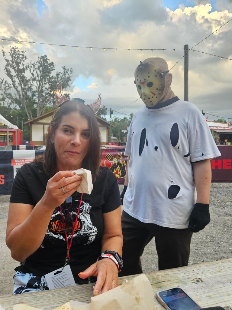 A woman eating a snack at an outdoor setting with a scare actor dressed in a horror-themed costume standing behind her.