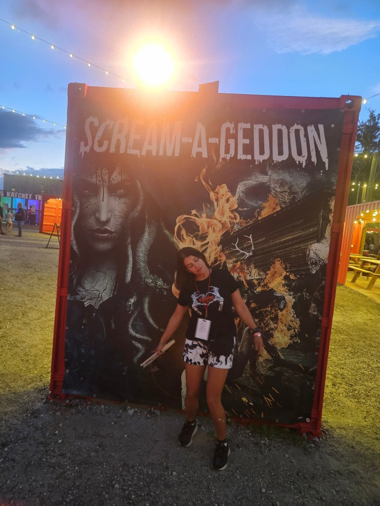 A person poses in front of a bright and colorful Scream-A-Geddon banner featuring a spooky design, surrounded by string lights and dusk scenery.