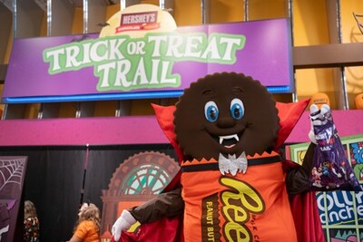 A festive scene at Hershey's Chocolate World featuring a mascot dressed as a chocolate cookie with a cape, holding a candy bag in front of a colorful 'Trick or Treat Trail' sign.